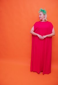 Φωτεινό ροζ, αμάνικο φόρεμα με ρέουσα σχεδίαση, που φτάνει στους αστραγάλους. Πράσινο αξεσουάρ κεφαλής με λεπτομέρειες από τούλι. Ζωντανό πορτοκαλί φόντο.