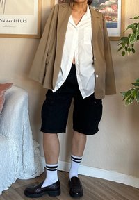 Χακί σακάκι πάνω από λευκό πουκάμισο με κουμπιά, μαύρες cargo σορτς, μαύρους loafers και λευκές κάλτσες με μαύρες ρίγες. Εσωτερικός χώρος με φυτά.