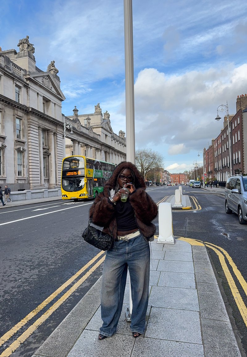 Woman in a fur jacket and wide-legged jeans drinking from a cup, standing on a pavement near a street with a yellow double-decker bus and historic buildings.