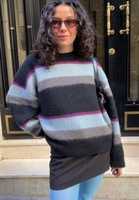 Γυναίκα με σγουρά μαλλιά που φοράει γυαλιά ηλίου, ριγέ αφράτο πουλόβερ, μαύρη φούστα και γαλάζια κολάν, στέκεται μπροστά από μαύρες πόρτες.