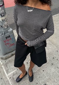 Svart og hvit stripete av-shoulder topp, svarte shorts og flate svarte sko; har også en liten svart veske med et teksturert mønster.