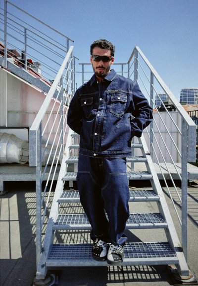 Chaqueta de mezclilla con dos bolsillos en el pecho y botones, combinada con pantalones de mezclilla de corte holgado. Calzado con un patrón en blanco y negro. Escaleras de metal gris.