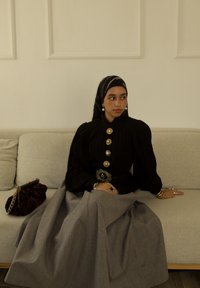 Blouse noire plissée avec de gros boutons et des épaules structurées, associée à une jupe grise. Les accessoires comprennent un collier de perles et un foulard à motifs.