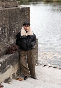 Persoon die een zwarte leren jas draagt met een grote beige schapenvachtkraag, bruine wijde broek en zwarte schoenen, leunend tegen een stenen muur bij het water.