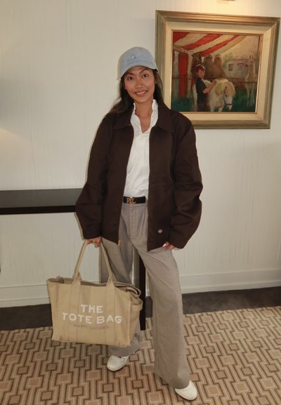Chaqueta oversized marrón, camisa blanca de botones, pantalones de cuadros grises de pierna ancha, bolsa tote beige etiquetada "THE TOTE BAG", gorra gris, zapatillas blancas.