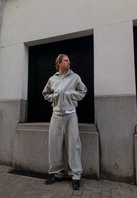 Un homme en sweat à capuche gris et pantalon de survêtement se tient les mains dans les poches contre un mur de bâtiment avec un volet noir fermé.