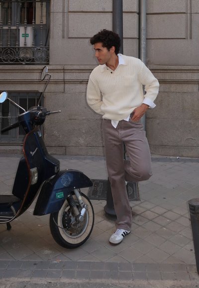 Suéter de punto en crema con un patrón texturizado, pantalones de pana marrones y zapatillas blancas. Scooter vintage negro al lado de un muro de piedra en la calle.