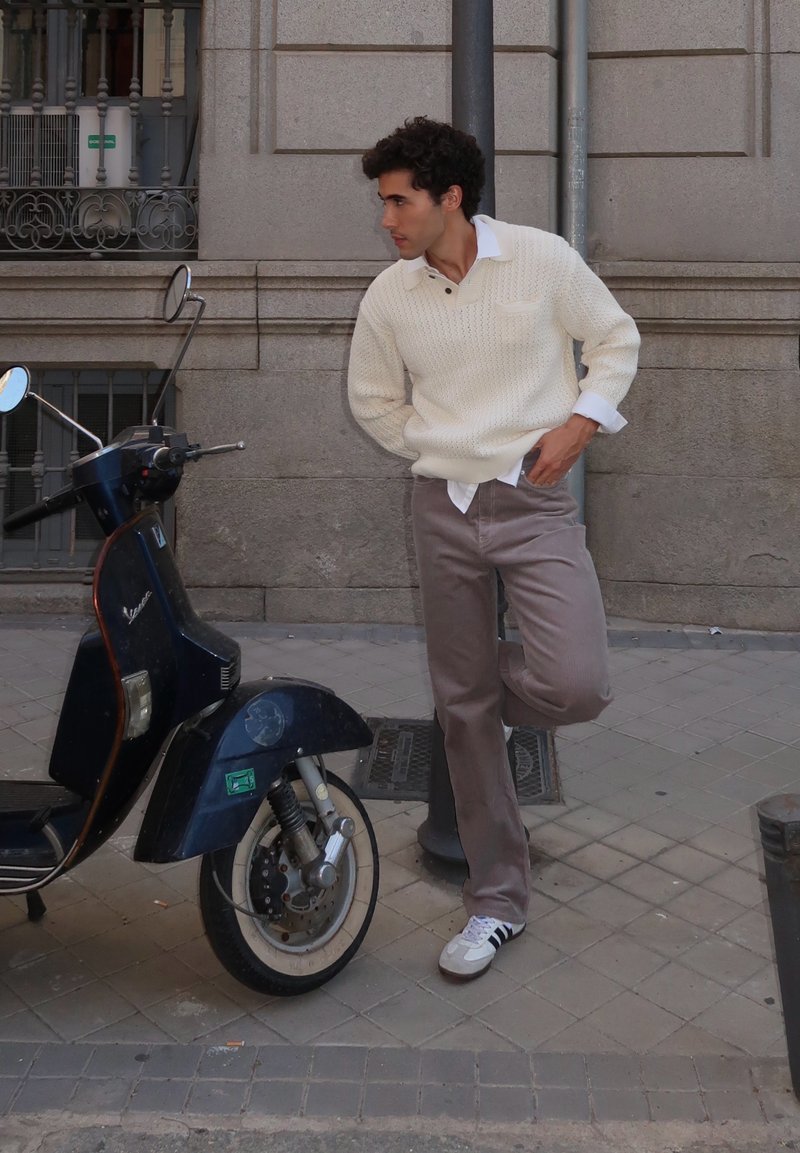 Kudum cream'ist kampsunist koos tekstuuriga mustriga, pruunist corduroy'ist pükstest ja valgetest tossudest. Must vintage skuter, mis on paigutatud kiviseina äärde tänaval.