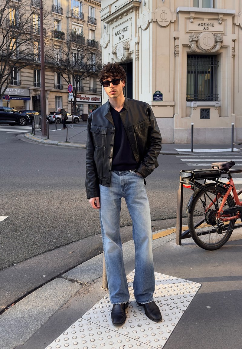 Young man in black leather jacket, sunglasses, and light blue jeans standing on a city street corner near a parked bicycle.