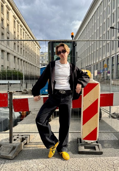 Joven con gafas de sol, camiseta blanca, chaqueta negra y zapatillas amarillas apoyado en barreras de construcción en un entorno urbano.