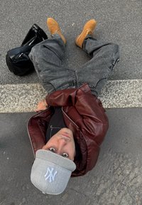 Giacca di pelle marrone, t-shirt nera, berretto da baseball grigio con logo, jeans grigi, stivali beige, seduto sul marciapiede accanto a una borsa nera.