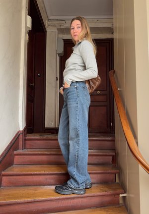 Joven mujer con camiseta gris de manga larga, vaqueros azules y zapatos negros de pie en una escalera de madera dentro de un edificio con puertas de madera oscura.