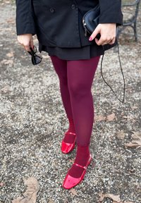 Κόκκινα παπούτσια από λακαρισμένο δέρμα με κο scalloped, φορεμένα με μ πορντό καλσόν και μαύρη φούστα, κρατούσαν γυαλιά και μια μικρή μαύρη τσάντα.