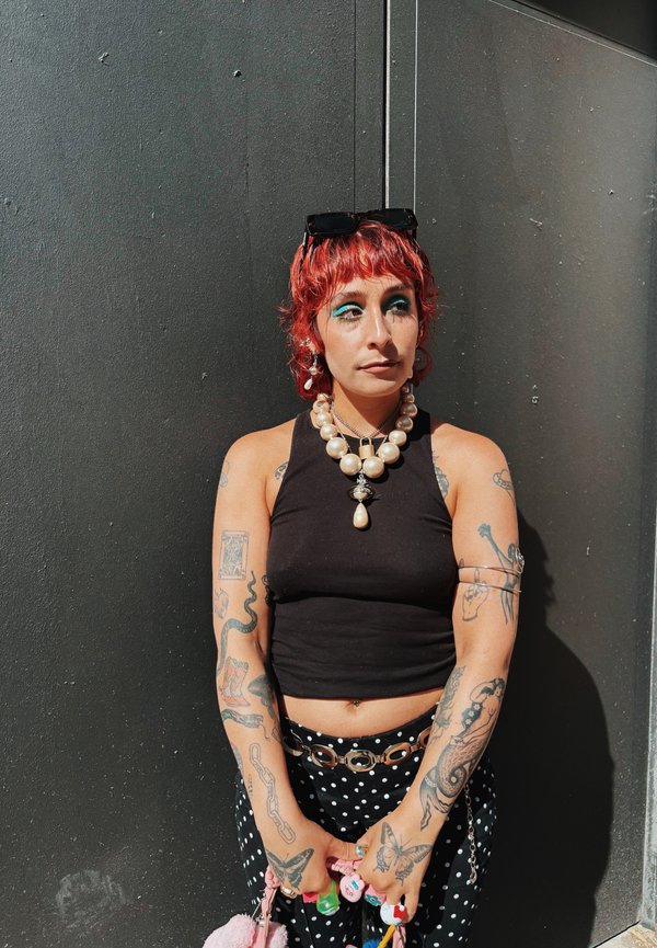 Kvinde med rødt hår og blå øjenmakeup står mod en mørk væg iført en sort tanktop, perlehalskæde, bukser med polkaprikker og holder farverige accessories.