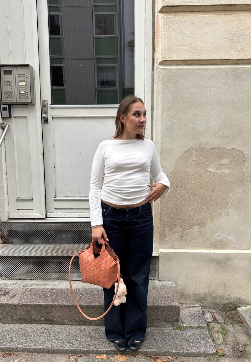 Bolsa de pêssego trançada com um brinquedo de pelúcia destacável, blusa de manga longa branca e jeans escuros de perna larga, contra um parede texturada.