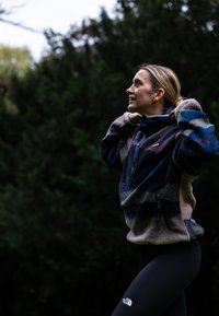 Fleeceová mikina s barevnými horizontálními pruhy, vybavená čtvrtinovým zipem, dekorativními knoflíky a boční kapsou na zip, kombinovaná s černými legínami.