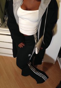 Persoană îmbrăcată cu un maiou alb, pantaloni de trening negri Adidas cu dungi albe, o haină neagră și ținând o poșetă bej.