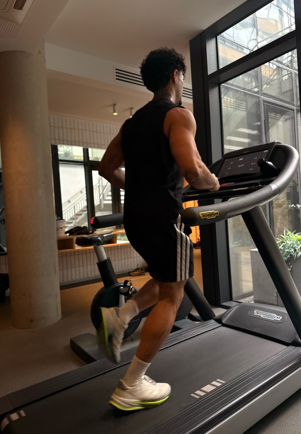 Άνδρας με μαύρη αθλητική ενδυμασία τρέχει σε διάδρομο γυμναστικής σε μοντέρνο γυμναστήριο με μεγάλα παράθυρα και στατικό ποδήλατο κοντά.