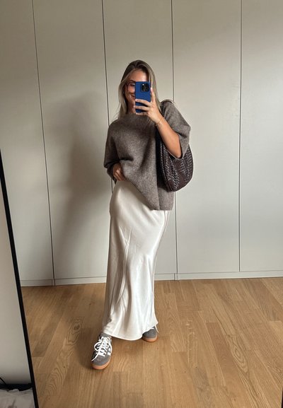 Suéter marrón de lana de gran tamaño con mangas cortas, combinado con una falda blanca de satén y zapatillas grises. La modelo sostiene un bolso de tejido oscuro.
