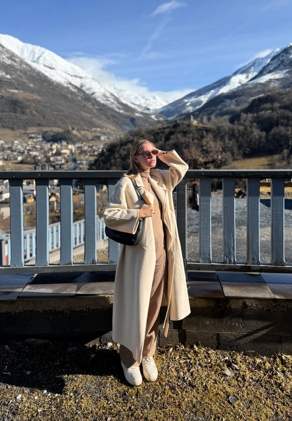 Femme en manteau beige et lunettes de soleil debout près d'une rambarde, avec des montagnes enneigées et un village en arrière-plan par une journée ensoleillée.