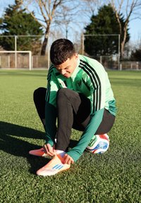 Jeune joueur de football masculin portant une veste verte, attachant des crampons orange tout en étant assis sur un ballon de football sur un terrain d'herbe.