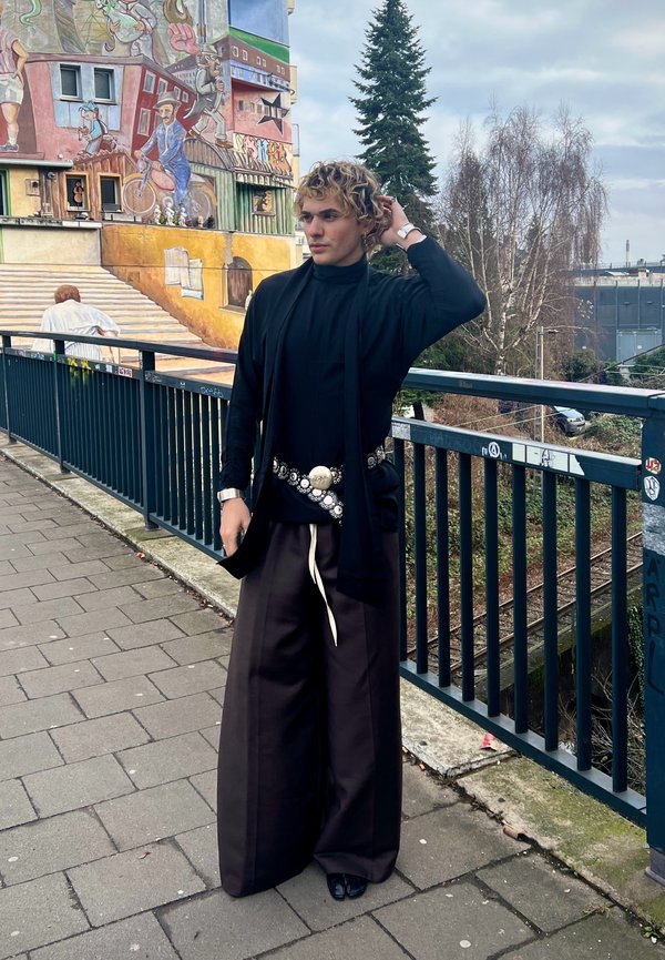 Άτομο με σγουρά μαλλιά που φοράει φαρδιά μαύρη παντελόνα, μαύρο τοπ και διακοσμητική ζώνη, στέκεται δίπλα σε κιγκλίδωμα με πολύχρωμο γκράφιτι και δέντρα στο παρασκήνιο.