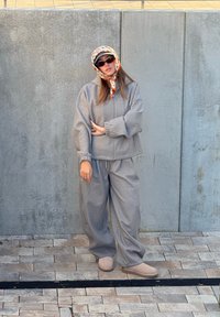 Femme portant une veste et un pantalon gris surdimensionnés, des chaussures beiges, un foulard à motifs, des lunettes de soleil, debout contre un mur en béton sur un sol carrelé.