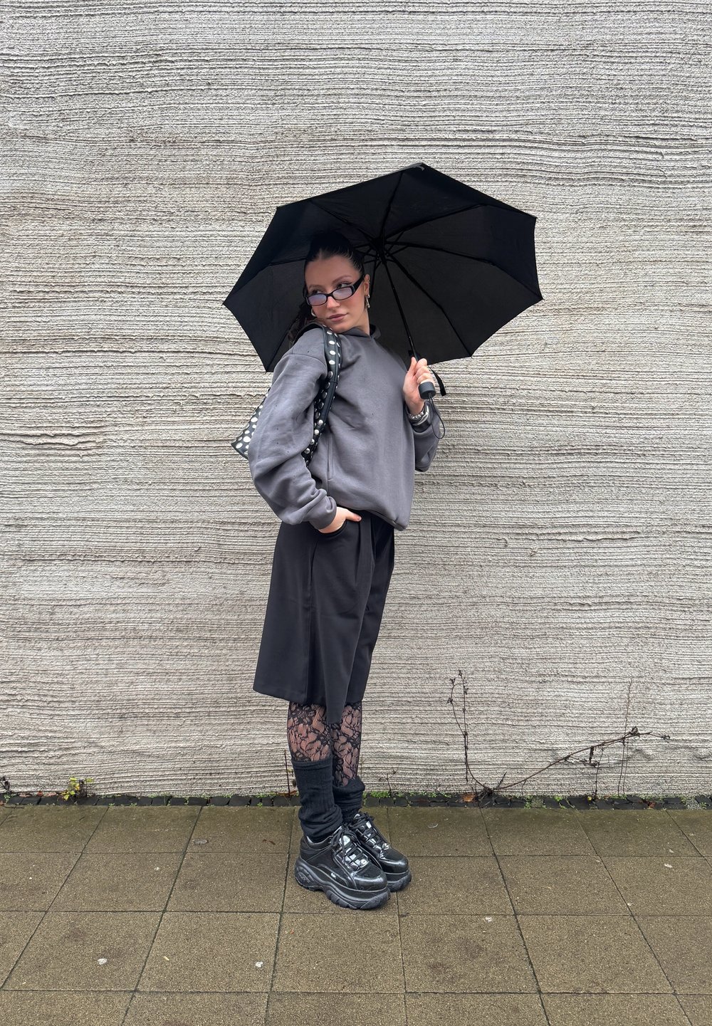 Musta sateenvarjo, jota pitää henkilö, joka on pukeutunut harmaaseen huppariin, mustiin shortseihin ja kuviollisiin legginsseihin, jalassaan mustat lenkkarit, taustalla teksturoitu seinä.