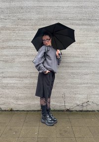 Musta sateenvarjo, jota pitää henkilö, joka on pukeutunut harmaaseen huppariin, mustiin shortseihin ja kuviollisiin legginsseihin, jalassaan mustat lenkkarit, taustalla teksturoitu seinä.