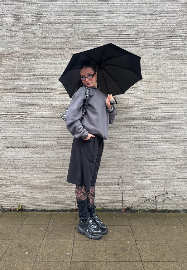 Musta sateenvarjo, jota pitää henkilö, joka on pukeutunut harmaaseen huppariin, mustiin shortseihin ja kuviollisiin legginsseihin, jalassaan mustat lenkkarit, taustalla teksturoitu seinä.