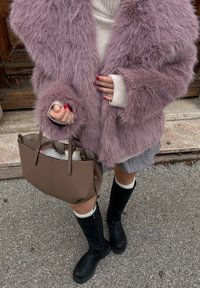 Personne portant un manteau rose duveteux, une jupe grise, un pull blanc, des bottes noires montantes jusqu'aux genoux, tenant un sac à main marron sur une surface en pierre et asphalte.