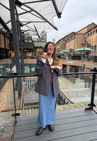 Mujer con gafas de sol, blazer gris y falda vaquera comiendo comida de una caja para llevar en un balcón cubierto al aire libre en una zona de mercado.