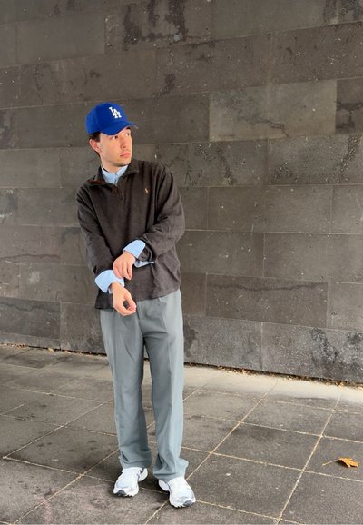 Hombre con gorra azul de Los Ángeles, suéter oscuro, camisa azul clara, pantalones grises y zapatillas blancas, de pie sobre un pavimento de baldosas frente a una pared de piedra oscura.