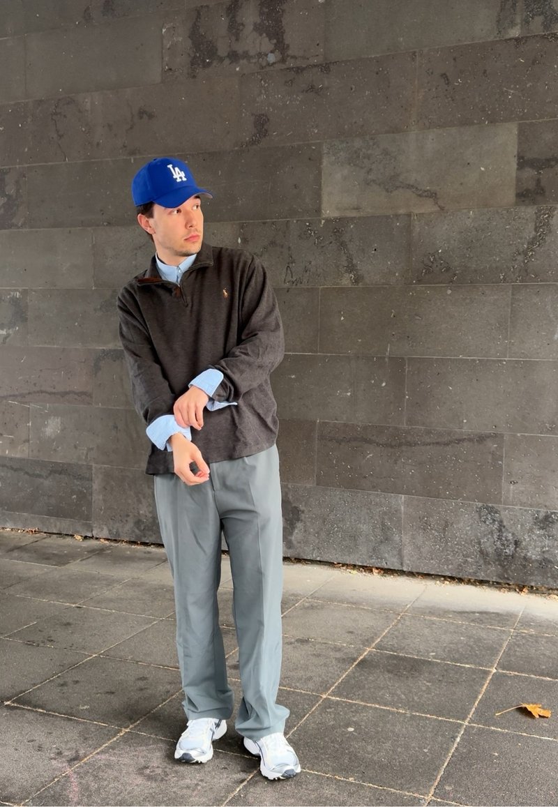 Homme portant une casquette bleue de Los Angeles, un sweatshirt sombre, une chemise bleu clair, un pantalon gris et des baskets blanches, se tenant sur un pavé carrelé contre un mur en pierre sombre.