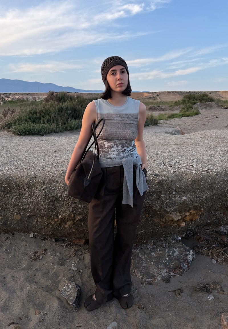 Jeune femme portant un top sans manches, un pantalon foncé et un bonnet en tricot, debout sur un terrain rocheux avec un grand sac à main et un pull attaché à la taille.