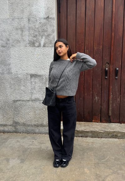 Mujer con suéter gris y pantalones negros, de pie con la mano cerca del cuello, con bolso negro cruzado, frente a una puerta de madera y una pared de piedra.