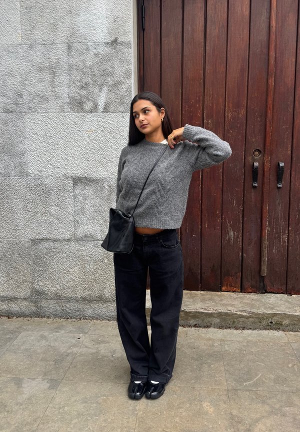 Kvinne i grå genser og svarte bukser står med hånden nær nakken, svart skulderveske, foran en tredør og en steinvegg.