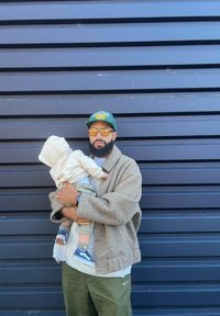 Ένας άνδρας με ένα καφέ πουλόβερ σε oversized στυλ κρατάει ένα παιδί με καφέ φούτερ. Και οι δύο φοράνε αθλητικά παπούτσια, φαίνεται μια μπλε τοίχος στο φόντο.