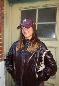 Bruine faux leren zip-up jas met een hoge kraag en zilveren hardware accenten. Bordeaurode pet met logo. Lichte houten achtergrond.
