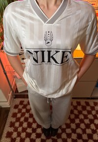 Maillot Nike rayé blanc avec un col en V gris, arborant un grand logo noir et le texte "Depuis 1972". Associé à un pantalon de jogging gris.