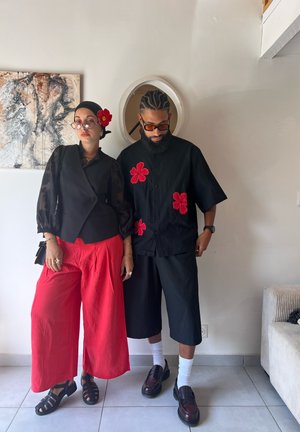 Dos individuos están uno al lado del otro. Uno lleva una blusa negra transparente de manga larga, un chaleco negro y pantalones rojos de pierna ancha. El otro lleva una camisa negra de manga corta con detalles florales rojos y pantalones cortos negros. Ambos tienen zapatos y accesorios únicos.