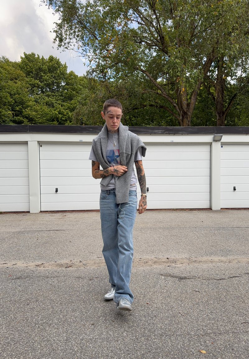 Personne aux cheveux courts et tatouages marche sur le trottoir, portant un T-shirt gris, un jean, des baskets et un pull gris drapé sur les épaules près des portes de garage.