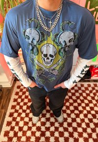 Personne portant un tee-shirt à manches longues superposées avec un motif crâne et serpent, des colliers en forme de croix, un jean foncé et des baskets grises, debout sur un tapis à carreaux.