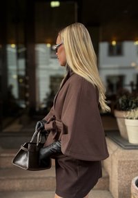 Ξανθιά γυναίκα με καφέ κάπα και μαύρα γάντια κρατά καφέ τσάντα, στέκεται πλάγια κοντά σε σκαλοπάτια και γλαστράκια έξω από ένα κτίριο.