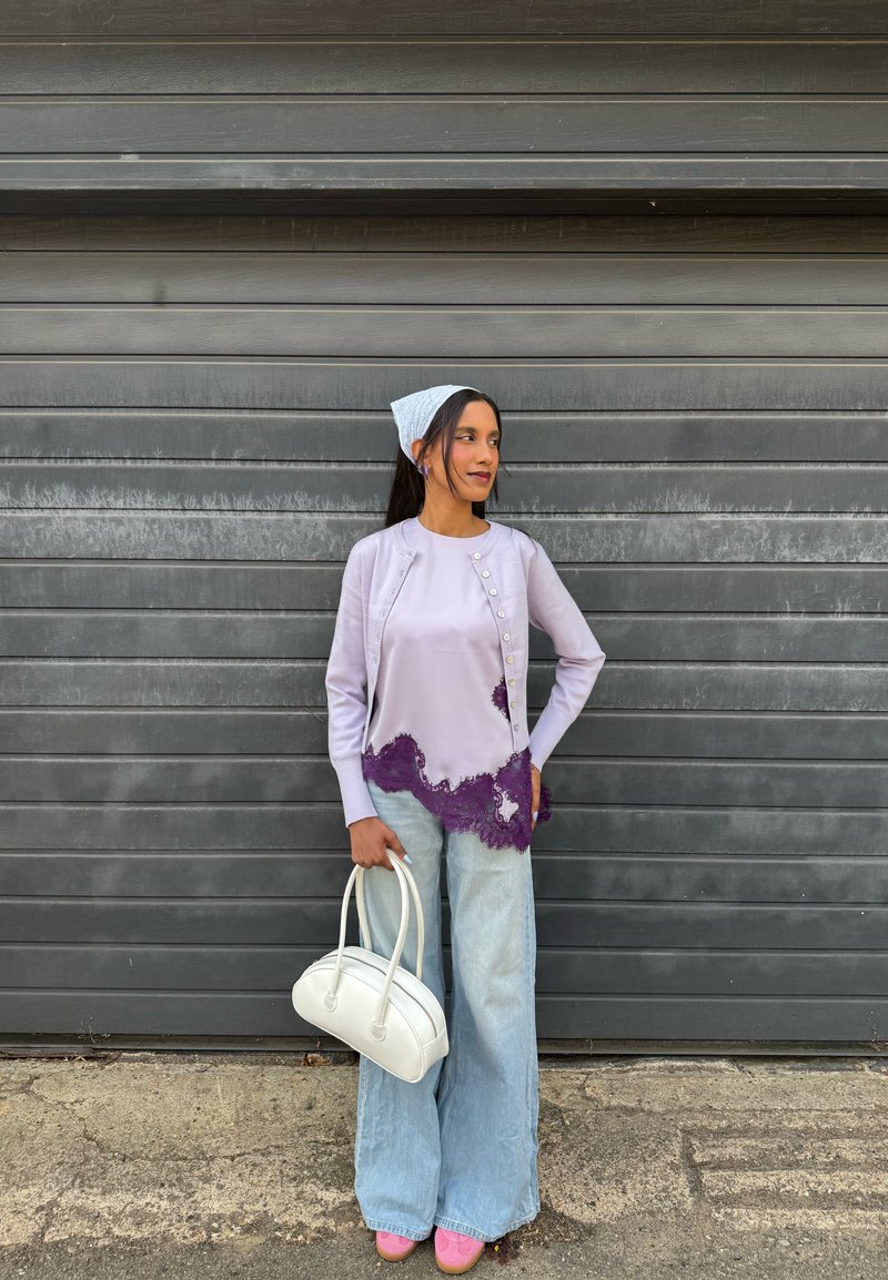 Femme debout contre un mur à volets gris portant un haut lavande avec de la dentelle violette, un jean large couleur délavé clair, un foulard blanc sur la tête et tenant un sac à main blanc.