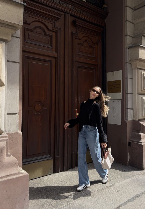Femme avec des lunettes de soleil et les cheveux longs, portant un haut noir et un jean, marche en tenant un sac à main blanc devant de grandes portes en bois sculpté.