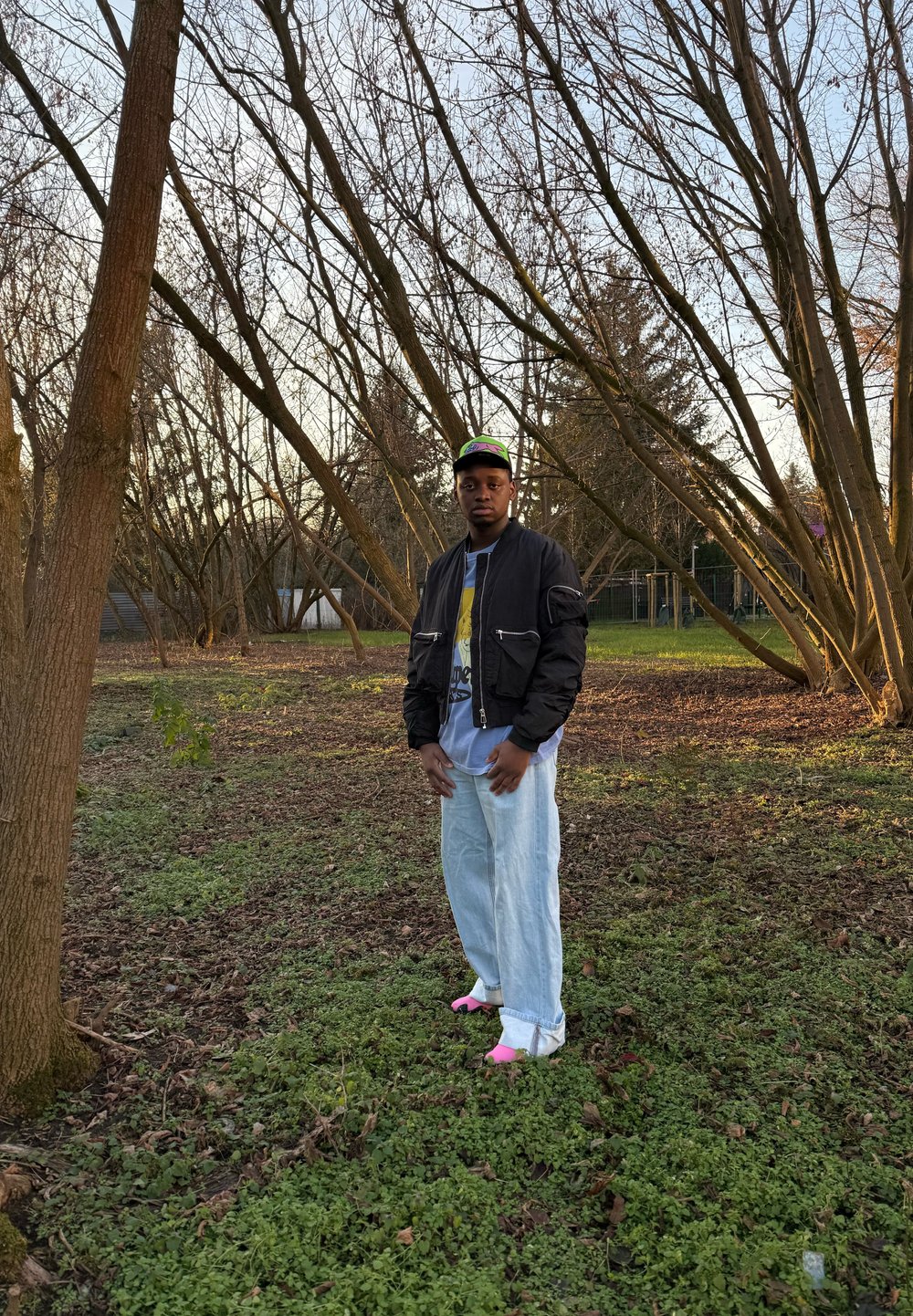 Fekete bomberdzseki cipzáras zsebekkel, grafikus póló, világoskék, bőszárú farmer, rózsaszín lábbeli egy erdős területen.