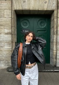 Joven con cabello rizado que lleva una chaqueta de cuero negra, pantalones blancos y un bolso marrón, de pie ante una gran puerta verde.