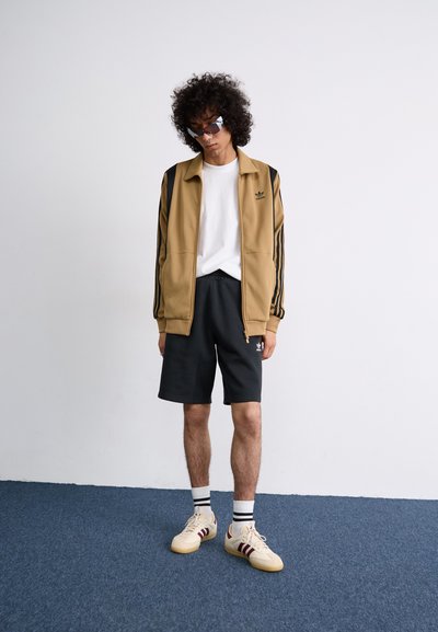 Chaqueta marrón con cremallera y rayas negras, camiseta blanca, pantalones cortos negros y zapatillas color beige con rayas rojas. El modelo también lleva gafas de sol espejadas.