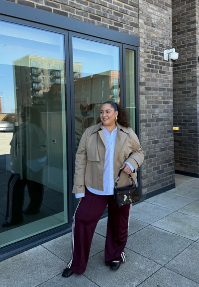 Chaqueta oversized beige, camisa blanca y pantalones burdeos con rayas blancas. Bolsa negra con estampado de vaca. De pie cerca de ventanas de cristal.